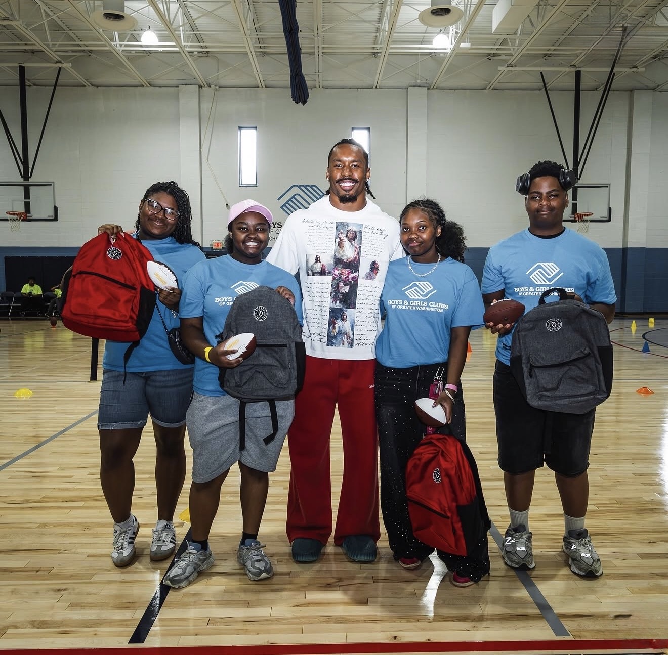 Nick Cross with Boys & Girls Clubs kids holding new backpacks and footballs in a gym
